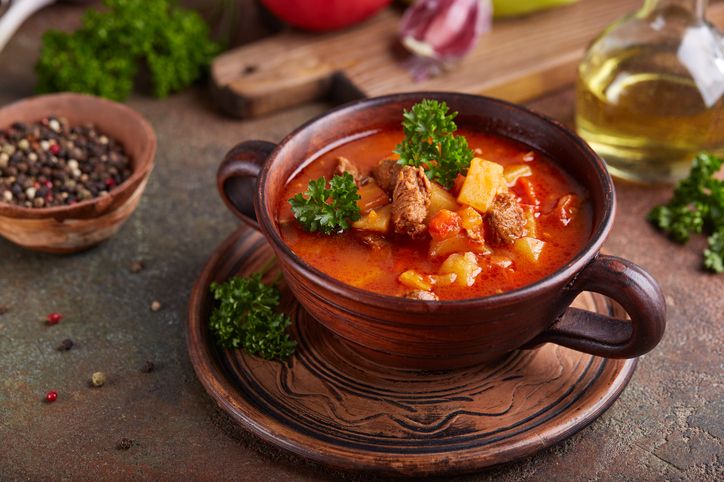 Gulaschsuppe in einer braunen Schale mit Fleisch, Kartoffeln, Gemüse und Petersilie, daneben Gewürze und Öl.