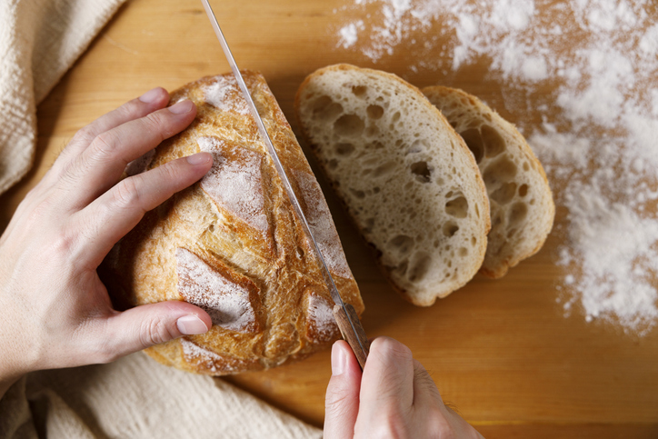 Sauerteigbrot wird auf einem bemehlten Holzbrett mit einem Brotmesser in dicke Scheiben geschnitten.