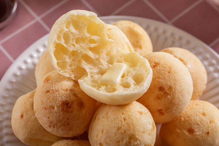 Aufgeschnittenes Pão de Queijo mit luftiger, lockerer Krume und geschmolzener Butter auf einem Teller mit weiteren Käsebrötchen.