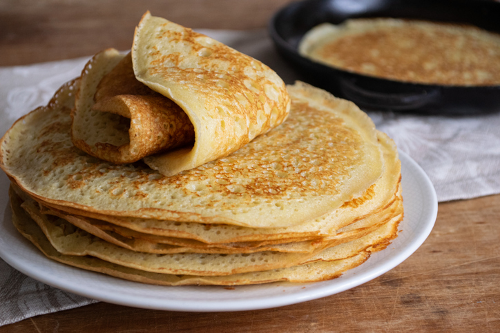 Goldbraun gebackene Crêpes auf einem weißen Teller mit einem aufgerollten Crêpe und einer Pfanne im Hintergrund.