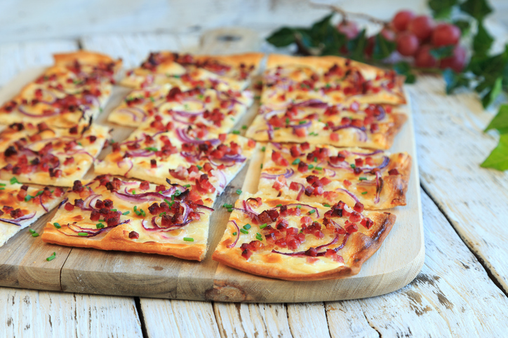 Mehrere Stücke Flammkuchen mit Crème fraîche, roten Zwiebeln, Speckwürfeln und Schnittlauch auf einem Holzbrett.
