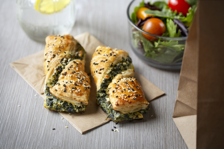 Vier Blätterteigtaschen mit Spinat-Käse-Füllung und Sesam auf einer Serviette, daneben eine Schüssel mit gemischtem Salat.