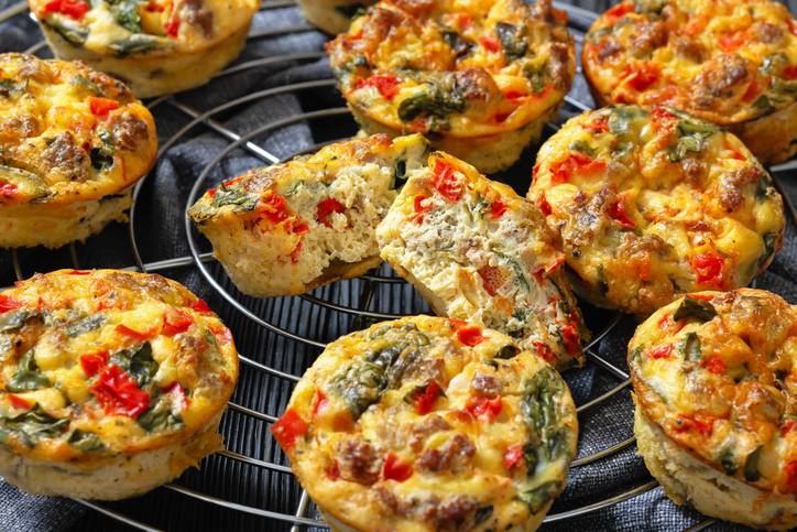 Mehrere Frittata Eier-Muffins auf einem Kuchengitter, darunter ein halbierter Muffin mit sichtbarer Gemüsefüllung.