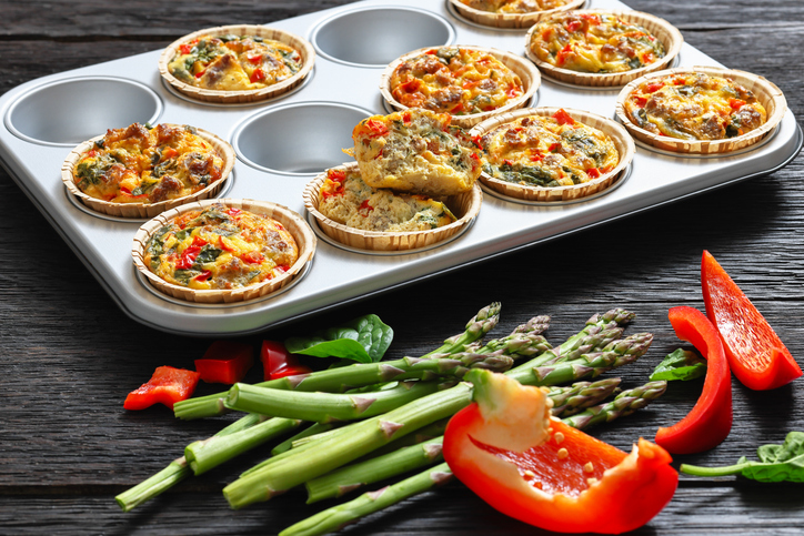 Frittata Eier-Muffins in einer Muffinform, dazu grüner Spargel und rote Paprikastücke auf einem dunklen Tisch.