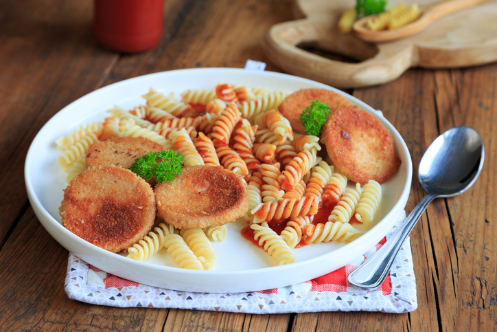 Jägerschnitzel mit Spirelli, fruchtiger Tomatensoße, panierten Jagdwurstscheiben und frischer Petersilie auf einem weißen Teller.
