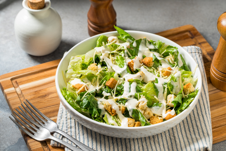 Schüssel mit Caesar Salad aus Römersalat, Croutons, Käse und cremigem Dressing auf einem Holzbrett