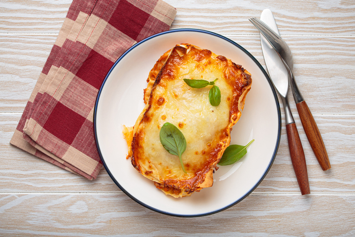 Einzelnes Stück Lasagne mit goldbraun geschmolzenem Käse und frischen Basilikumblättern auf einem weißen Teller.
