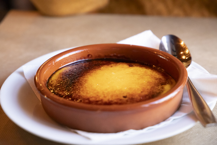 Crema Catalana in einer braunen Tonschale mit goldbrauner Karamellschicht auf einem weißen Teller mit Löffel