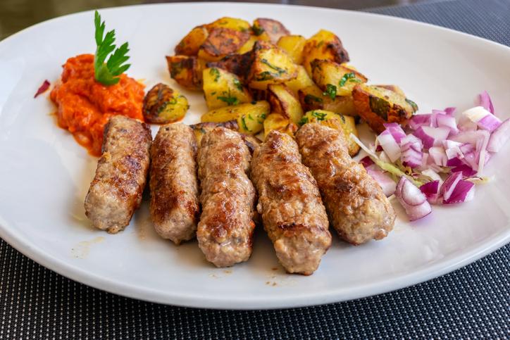 Gegrillte Cevapcici auf einem weißen Teller mit Bratkartoffeln, Ajvar und gehackten roten Zwiebeln.