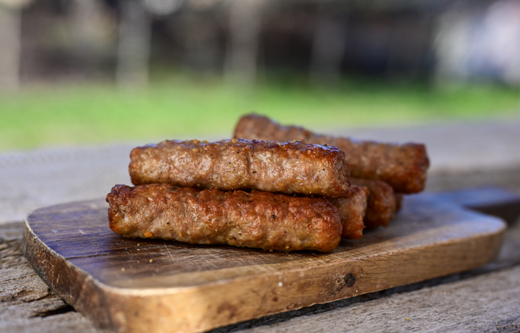 Mehrere gegrillte Cevapcici liegen auf einem rustikalen Holzbrett im Freien übereinandergestapelt.