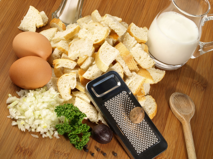 Zutaten für Semmelknödel mit Brotwürfeln, Eiern, Milch, Zwiebeln, Petersilie und Muskatnuss auf einer Holzfläche.