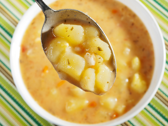 Löffel mit Kartoffelstücken über einer Schüssel cremiger Kartoffelsuppe mit sichtbaren Gemüsestückchen.