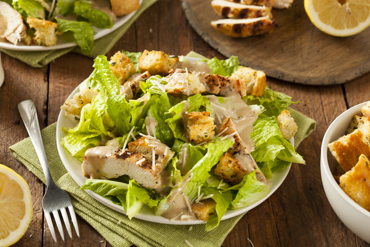 Teller mit Caesar Salad, knusprigen Croutons, Hähnchenstücken und Dressing auf einem Holztisch