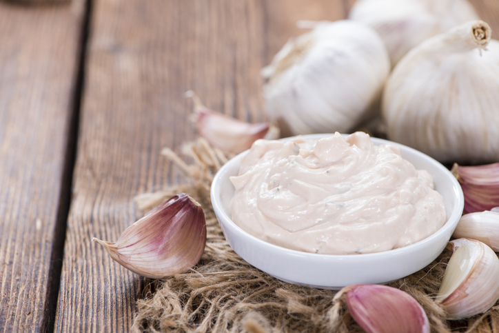 Helle Aioli in einer weißen Schale auf rustikalem Holzuntergrund, umgeben von ganzen und einzelnen Knoblauchzehen.