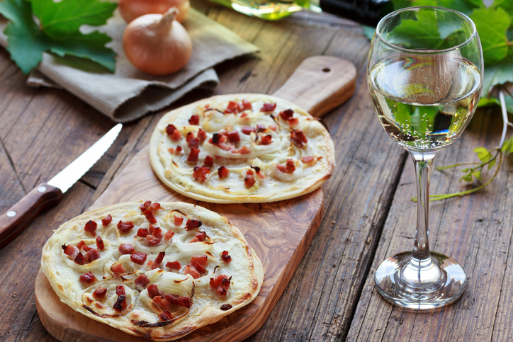 Zwei kleine Flammkuchen mit Crème fraîche, Zwiebeln und Speck auf einem langen Holzbrett neben einem Glas Weißwein.