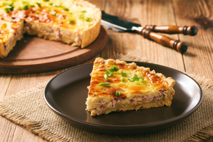 Stück Quiche Lorraine mit Speck, cremiger Eierfüllung, Käse und Frühlingszwiebeln auf einem dunklen Teller.