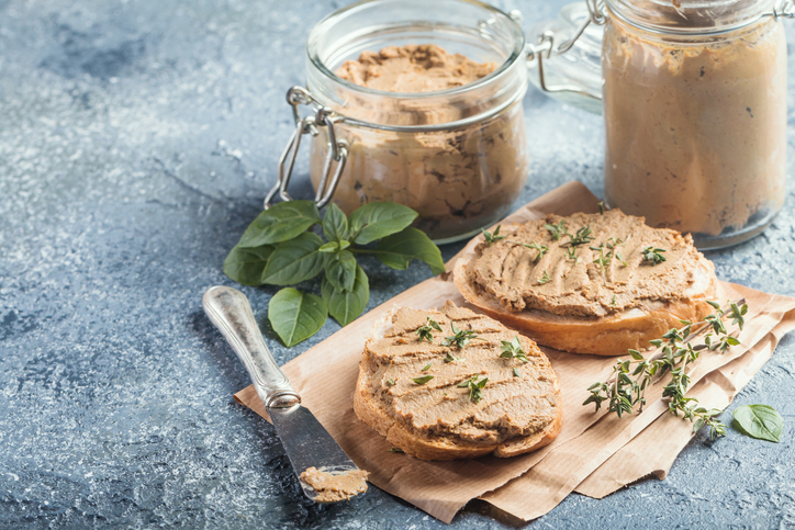 Hähnchenleber-Pastete auf zwei Brotscheiben mit Kräutern, daneben Gläser mit Pastete auf einer hellen Unterlage.