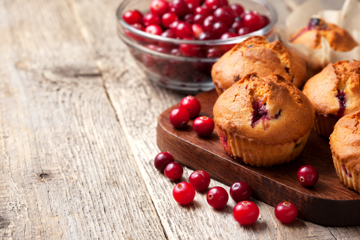 Frisch gebackene Cranberry Muffins auf einem Holzbrett mit roten Cranberries und einer Schale voller frischer Beeren im Hintergrund.