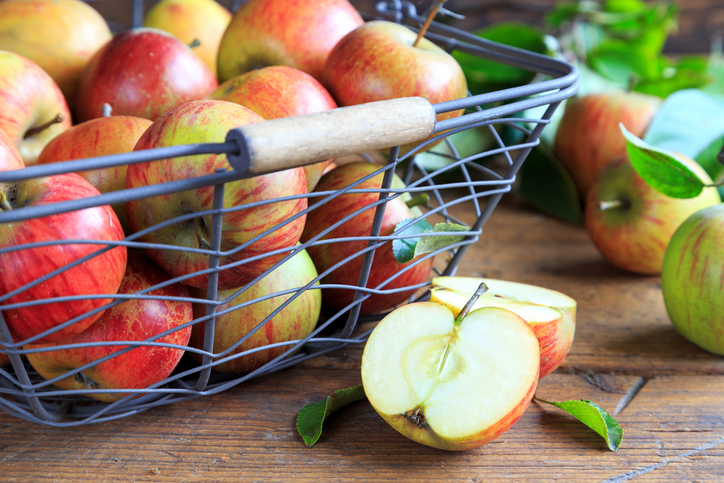 Rot-gelbe Äpfel in einem Metallkorb auf einem Holztisch, daneben ein halbierter Apfel und weitere Früchte mit grünen Blättern.