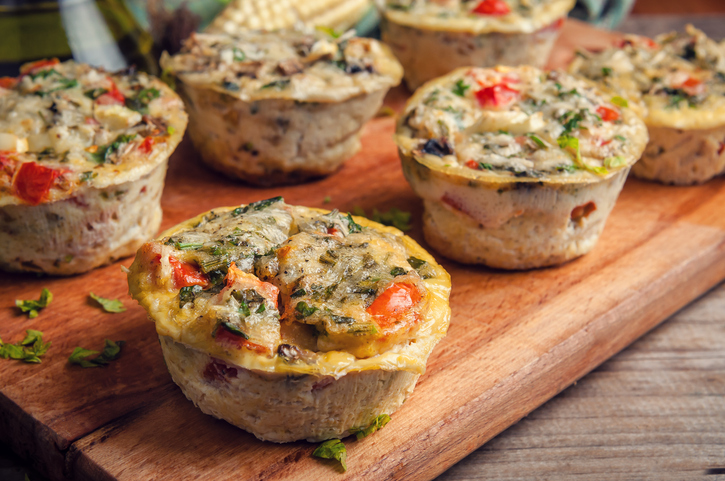 Mehrere Frittata Eier-Muffins mit Gemüse und Kräutern auf einem Holzbrett, vorne ist ein Muffin aufgeschnitten.