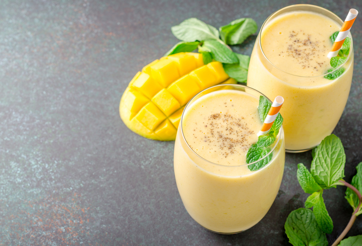 Zwei Gläser Mango Lassi mit frischer Minze und Trinkhalmen, daneben eine aufgeschnittene Mango auf dunklem Untergrund.