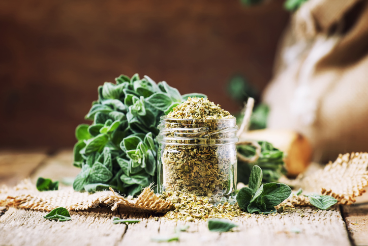 Getrockneter Oregano in einem kleinen Glas, umgeben von frischen grünen Oreganoblättern auf einem rustikalen Holztisch.