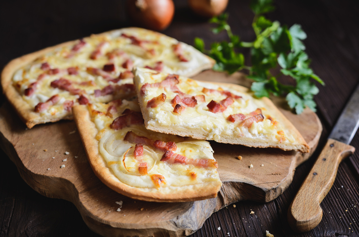 Flammkuchen auf einem Holzbrett mit heller Creme, Zwiebelringen und gebratenen Speckwürfeln, teilweise in Stücke geschnitten.
