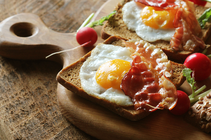 Strammer Max mit Spiegelei und knusprigem Speck auf dunklem Brot, angerichtet mit frischen Radieschen auf einem Holzbrett.