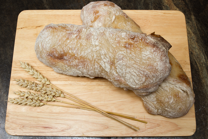 Zwei rustikale Ciabatta Brote mit heller bemehlter Kruste auf einem Holzbrett neben mehreren Weizenähren.