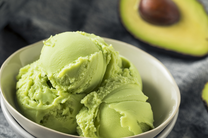 Mehrere Kugeln Avocado-Eis in einer hellen Schale, im Hintergrund ist eine halbierte Avocado zu sehen.