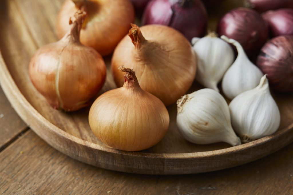 Mehrere gelbe Zwiebeln, rote Zwiebeln und Knoblauchknollen liegen nebeneinander auf einem runden Holztablett, das auf einem Holztisch steht.