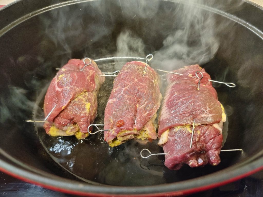 Drei gefüllte Rinderrouladen werden in einem Bräter scharf angebraten, um eine kräftige Kruste zu entwickeln.