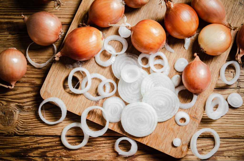 Mehrere braune Zwiebeln und in Ringe geschnittene weiße Zwiebelscheiben liegen auf einem Holzschneidebrett, umgeben von einer rustikalen Holztisch-Oberfläche.