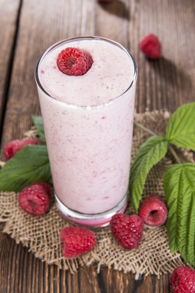 Ein Glas mit cremigem rosa Himbeer-Shake, garniert mit einer Himbeere, daneben frische Himbeeren und grüne Blätter auf rustikalem Holztisch.