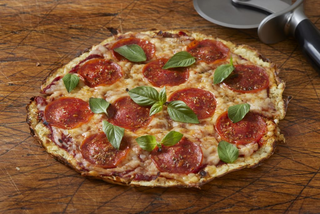Eine ganze Salamipizza mit geschmolzenem Käse, Peperoni-Salami und frischem Basilikum auf einem Holzbrett, daneben ein Pizzaschneider.
