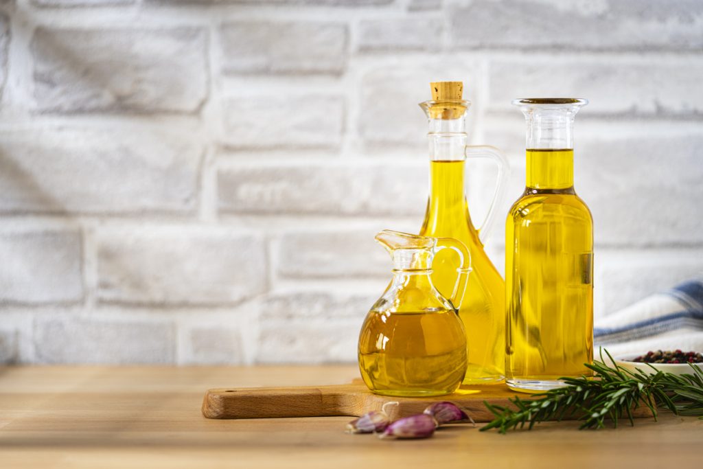 Mehrere Glasflaschen mit Speiseöl, arrangiert auf einem Holztisch mit Knoblauch und Rosmarin.