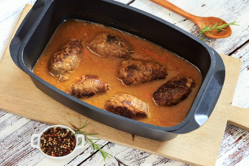 Traditionelle Rinderrouladen schmoren in einer dunklen Bratensoße in einem Bräter auf einem rustikalen Holztisch.