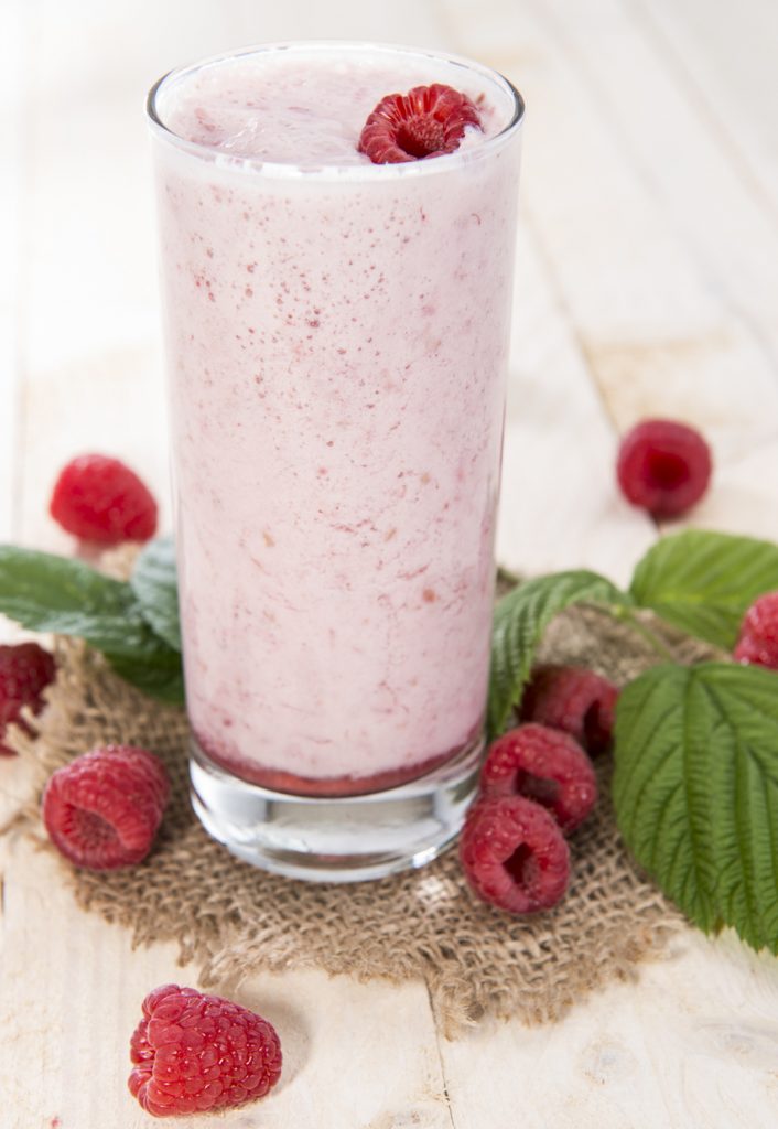 Ein hohes Glas mit cremigem rosa Himbeer-Shake, garniert mit einer Himbeere, umgeben von frischen Himbeeren und grünen Blättern auf hellem Holztisch.