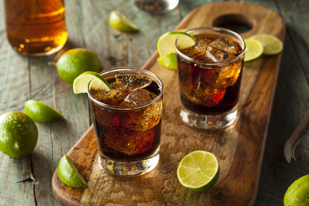 Zwei Gläser mit Cuba Libre, gefüllt mit Cola, Rum, Eiswürfeln und garniert mit Limettenscheiben, auf einem Holzbrett mit frischen Limetten.