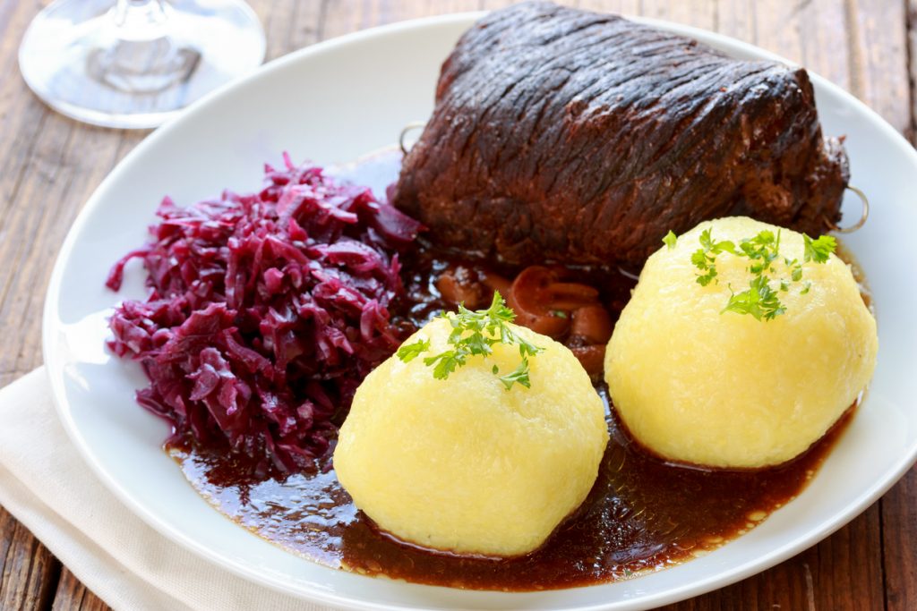 Traditionelles Gericht mit Rinderroulade in dunkler Bratensoße, serviert mit zwei Kartoffelklößen und Rotkohl auf einem weißen Teller.
