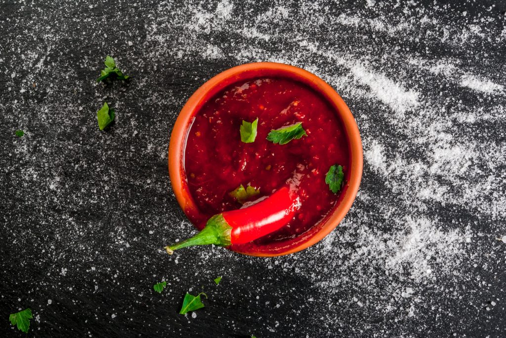 Schale mit roter Tabasco-Sauce, dekoriert mit frischer Chili und Kräutern, auf schwarzem Untergrund mit Salz.