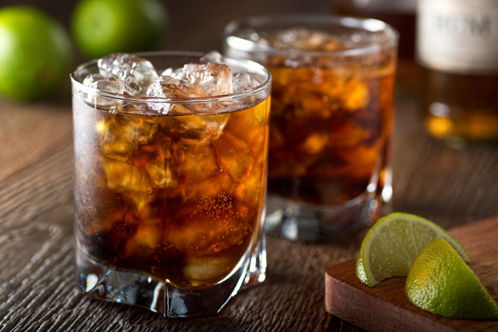 Zwei Gläser mit Cuba Libre, gefüllt mit Cola, Rum und Eiswürfeln, daneben Limettenstücke auf einem Holztisch.