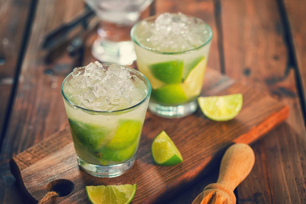 Zwei Gläser Caipirinha mit Limettenstücken und Crushed Ice, serviert auf einem Holzbrett mit frischen Limettenspalten und einem Holzstößel.