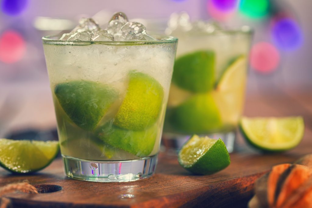 Zwei Gläser Caipirinha mit frischen Limettenstücken und Crushed Ice, serviert auf einem Holzbrett vor buntem, unscharfem Hintergrund.
