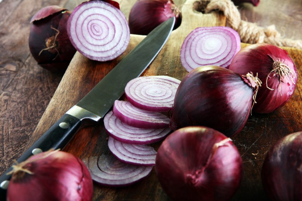 Mehrere rote Zwiebeln, teils im Ganzen und teils in Scheiben geschnitten, liegen auf einem hölzernen Schneidebrett neben einem scharfen Küchenmesser.