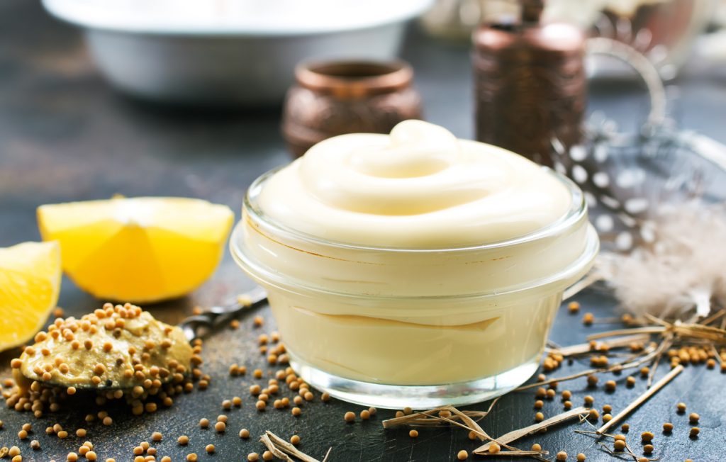 Ein Glas mit cremiger Mayonnaise, daneben Zitronenspalten, ein Löffel mit körnigem Senf und verstreute Senfkörner auf dunkler Oberfläche.
