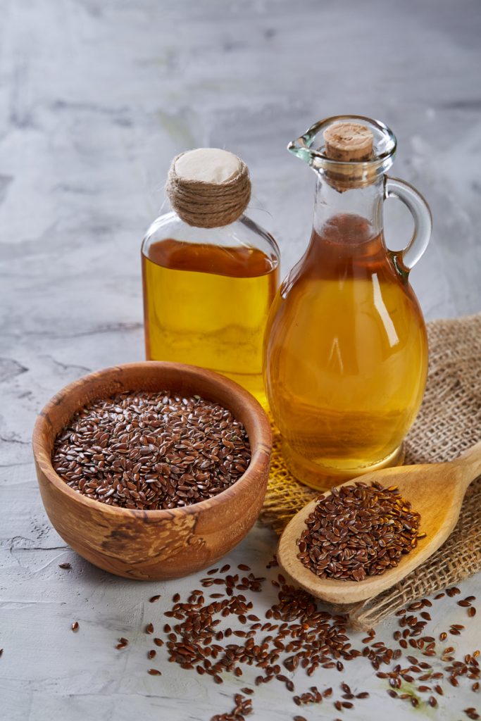 Zwei Glasflaschen mit goldenem Leinöl neben einer Holzschale und einem Holzlöffel voller brauner Leinsamen; einzelne Samen liegen verstreut auf hellem Untergrund.