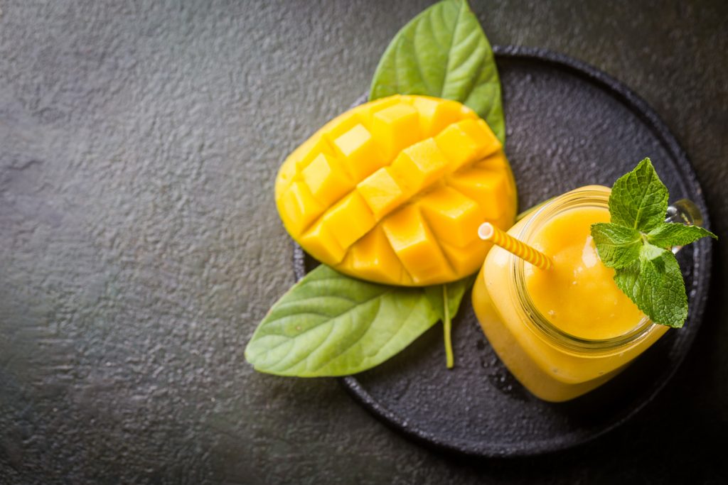 Mango-Smoothie im Glas mit Minze, daneben eingeschnittene Mango auf dunklem Teller