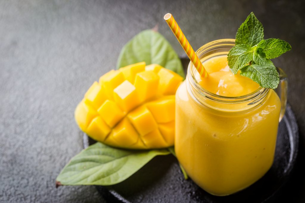 Mango-Smoothie im Glas mit Minze, daneben eingeschnittene Mango auf dunklem Teller