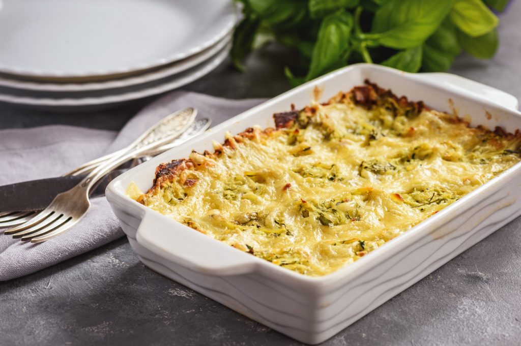 Zucchini-Kartoffel-Auflauf in weißer Form mit Käsekruste, daneben Teller und Besteck.